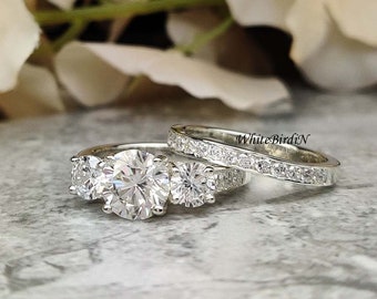 Peut inclure: Une bague de fiançailles en argent à trois pierres avec une grosse pierre centrale et deux pierres plus petites de chaque côté. La bague est associée à une bande argentée assortie avec de petites pierres serties en pavé. Les bagues sont sur une surface grise texturée.