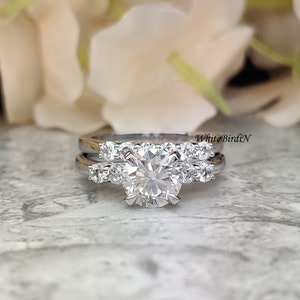 Puede incluir: Un conjunto de anillo de compromiso de plata con una piedra central redonda grande y piedras redondas más pequeñas en la banda. El anillo está sobre una superficie blanca.