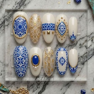 May include: A set of ten press-on nails featuring a white base with intricate blue tile-inspired patterns, gold metallic accents, gold chain motifs, and small white pearl embellishments, all arranged on a clear rectangular display stand.
