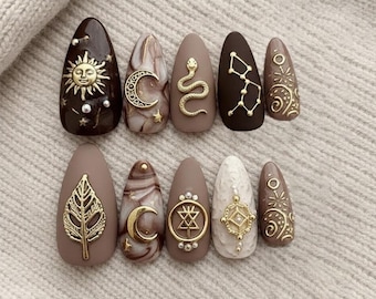 May include: A set of ten almond-shaped press-on nails in shades of dark brown, beige, and cream. The nails feature gold-toned metallic embellishments including a sun, crescent moon, snake, constellation, leaf, and geometric symbols with small pearls.