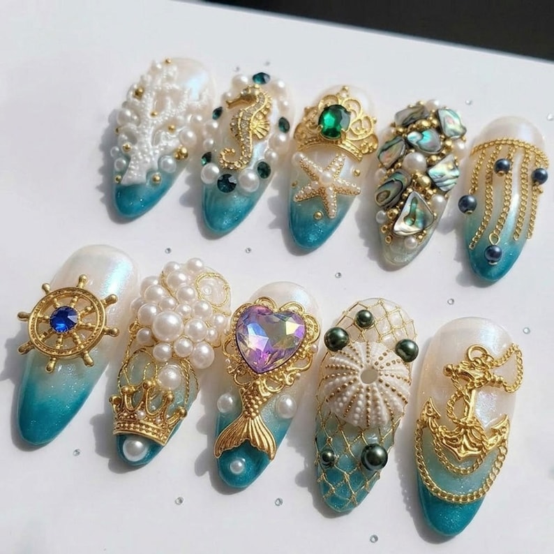 May include: A collection of ten intricately designed nail art pieces. Each nail features a white base with a gradient of turquoise at the tips. Embellishments include pearls, gold charms, and sea-themed designs like seahorses, starfish, and anchors.