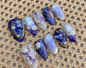 May include: An assortment of artificial nails in various designs. The nails feature a range of colors, including blue, purple, and white, with butterfly and floral embellishments. Some nails have glitter and pearl accents.