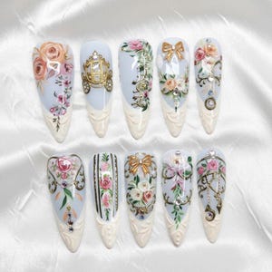 Puede incluir: Un juego de diez uñas postizas en forma de almendra con base azul claro, patrones florales detallados, detalles metálicos dorados, pequeños diamantes de imitación y adornos en 3D que incluyen una carroza, lazos y dijes colgantes.