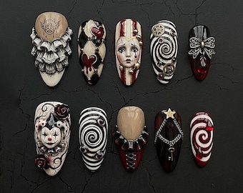May include: A set of ten almond-shaped press-on nails featuring intricate designs including a jester face, a sad clown, red hearts, black and white spirals, metallic gears, a bow with a red gem, a star with a chain and key, and ruffled collars.