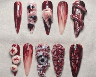 Pode incluir: Um conjunto de dez unhas postiças em formato stiletto com designs de terror. As unhas exibem padrões marmorizados em vermelho escuro e branco, dentes esculpidos em três dimensões, múltiplos globos oculares, um verme enrolado, texturas cerebrais e teias de aranha pretas.