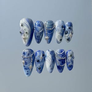 Puede incluir: Diez uñas postizas con diseño veteado azul y blanco. Cada uña está adornada con abalorios de estrella plateada, cuentas y delicadas cadenas. Las uñas, de forma ovalada, están dispuestas en dos filas de cinco.