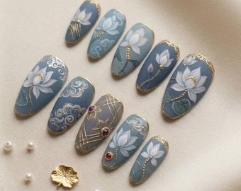 May include: A collection of ten oval-shaped artificial nails, each painted with a unique design. The nails feature a matte blue-gray base with white lotus flower designs, gold accents, and decorative embellishments. Some nails have gold borders and small gemstones.