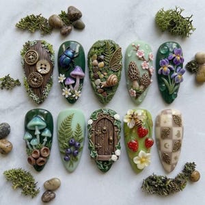 May include: Ten almond-shaped nail art tips featuring 3D sculpted designs including a wooden door, mushrooms, a beetle, a snail, strawberries, a bee, flowers, berries, and a checkered pattern with gold leaf accents, surrounded by moss and stones.