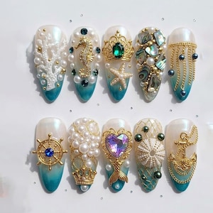 May include: A collection of ten oval-shaped, pearl-white artificial nails with a gradient teal base. Each nail is adorned with ocean-themed embellishments, including pearls, gold seahorses, crowns, and anchors. Some nails feature a coral design or a starfish.