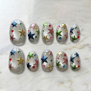 Puede incluir: Un conjunto de diez u&ntilde;as postizas ovaladas. Cada u&ntilde;a es transparente con un brillo nacarado y adornada con adornos en forma de estrella coloridas y peque&ntilde;as gemas redondas en varios colores. Las estrellas son en tonos rosa, azul, verde y dorado.
