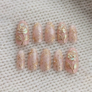 May include: A set of ten artificial nails in various sizes. Each nail is painted a light pink with gold floral designs. Some nails feature a rose design, while others have a floral pattern with small pearl accents. The nails are oval-shaped.