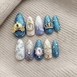 Puede incluir: Un conjunto de diez uñas artificiales con temática de cuento de hadas. Los diseños incluyen un carruaje dorado, un reloj, un tacón alto, una corona y detalles florales y de joyas. Las uñas son de varios tonos de azul, blanco y dorado con purpurina.