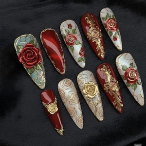 May include: A collection of ten artificial nails with various designs. The nails feature red, gold, and white colors, with rose and floral patterns. Some nails have a 3D rose design, while others have gold embellishments.