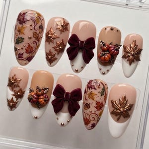 Puede incluir: Un juego de diez uñas postizas con temática otoñal sobre bases nude y blancas. Los diseños incluyen hojas de arce en bronce metálico, pequeñas calabazas naranjas y lazos de terciopelo burdeos, acentuados con tachuelas doradas y follaje pintado.