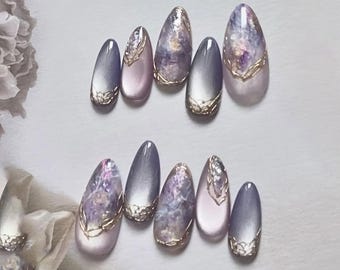 May include: A set of artificial nails in various shapes and sizes. The nails feature a gradient of colors, including lavender, pink, and gray, with gold trim and a marbled design. The nails are arranged on a white surface.