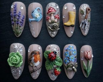 Puede incluir: Un juego de diez uñas postizas en forma de almendra con arte de jardín en 3D. Los diseños incluyen lavanda, una regadera azul, setas con un caracol, una bota amarilla, una horca de jardín, una col verde, un petirrojo, una mariquita y flores.