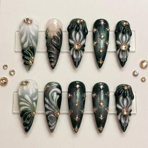May include: A set of ten long, pointed false nails. The nails feature a variety of designs, including white and green floral patterns, swirling gray and white designs, and dark green glitter. Each nail is adorned with small, gold-colored gems.