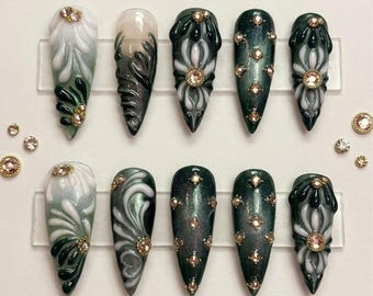 May include: A set of ten long, pointed false nails. The nails feature a variety of designs, including white and green floral patterns, swirling gray and white designs, and dark green glitter. Each nail is adorned with small, gold-colored gems.