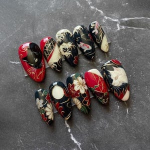May include: A collection of ten almond-shaped false nails with Japanese-inspired artwork. The nails display black, red, white, and gold patterns, including cats, flowers, and waves. The designs are detailed and artistic, combining traditional and modern elements.