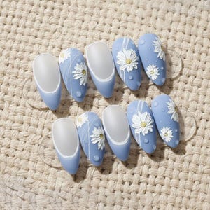 Puede incluir: Un conjunto de diez uñas artificiales en color azul claro, con acabado mate en las puntas. Cada uña presenta un diseño de margarita blanca con centros amarillos y detalles blancos. Las uñas están dispuestas sobre un expositor transparente.