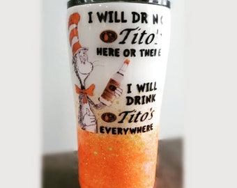 May include: A white tumbler with a black rim and an orange glitter base. The tumbler features a cartoon character holding a bottle and the text "I WILL DRINK Tito's HERE OR THERE I WILL DRINK Tito's EVERYWHERE."