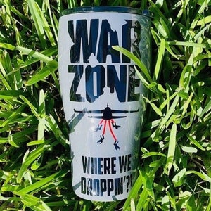May include: A white tumbler with the words "WAR ZONE" in large black letters. Below the text is an illustration of a plane with red lines and silhouettes of people. The phrase "WHERE WE DROPPIN?" is printed at the bottom.