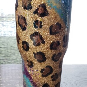 May include: A glitter-covered tumbler with a leopard print design. The tumbler features a gold glitter base with brown leopard spots. The top section has a blue and teal glitter design, and it has a black lid.