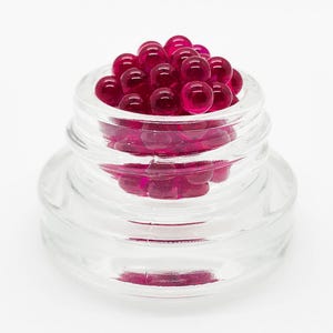 May include: A small, clear glass jar filled with numerous translucent, deep magenta spherical beads. The container features a threaded neck and a wide, circular base, showcasing the vibrant, glossy texture of the round, berry-colored contents.