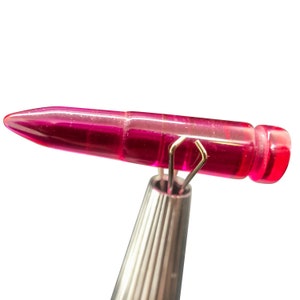May include: A translucent, vibrant magenta bullet-shaped object held by a pair of silver-toned metal tweezers. The object features a smooth, cylindrical body with a pointed tip and a flat, circular base, exhibiting a clear, glass-like appearance.
