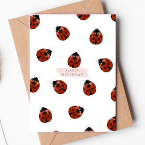 May include: A white rectangular greeting card featuring a repeating pattern of red and black ladybirds. The centre of the card displays a light pink horizontal brushstroke with the text HAPPY BIRTHDAY printed in small, dark, capitalised letters.