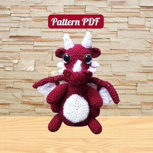 Puede incluir: Un dragón rojo de crochet con detalles blancos y grandes ojos negros. El dragón está sentado sobre una superficie de madera. El texto "Pattern PDF" se muestra sobre el dragón.