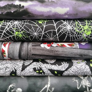 May include: A stack of five different fabric panels with Halloween themes. The top panel is a purple and white ombre with a dark silhouette of a forest. The second panel is black with white spiderwebs and green spiders. The third panel is a grey and white woodgrain pattern with a red and white candy cane. The fourth panel is black with white mummy figures. The bottom panel is grey with white skeletons.