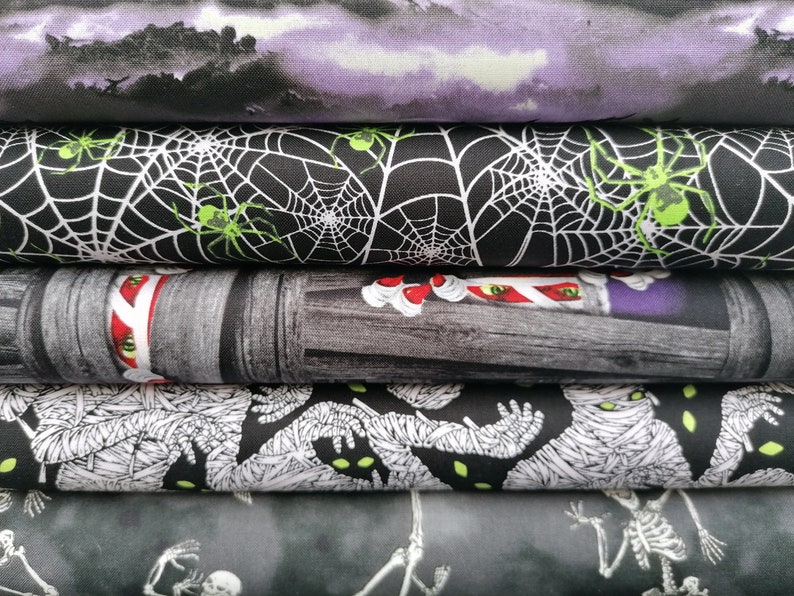 May include: A stack of five different fabric panels with Halloween themes. The top panel is a purple and white ombre with a dark silhouette of a forest. The second panel is black with white spiderwebs and green spiders. The third panel is a grey and white woodgrain pattern with a red and white candy cane. The fourth panel is black with white mummy figures. The bottom panel is grey with white skeletons.