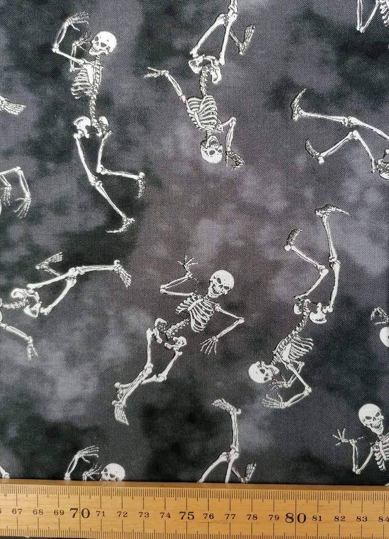 May include: Black and white fabric with a repeating pattern of skeletons dancing on a grey background. The fabric is measured with a ruler in inches.