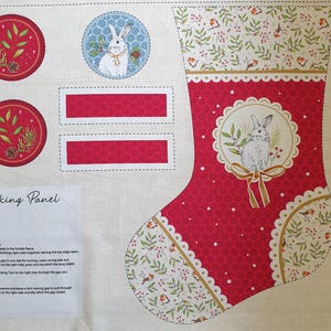 May include: A fabric sewing panel featuring a Christmas stocking design with a red background, white polka dots, and a central rabbit illustration. The panel includes two circular motifs with botanical prints, two rectangular strips, and printed instructions.