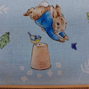 May include: Light blue fabric featuring a repeating pattern of a brown rabbit wearing a blue jacket jumping over a terracotta flower pot with a small blue bird perched on top, alongside green oak leaves, with a wooden ruler visible at the bottom.