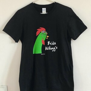 Może przedstawiać: Czarny T-shirt z zielonym kogutkiem w koronie z kwiatów i napisem "Frida Kellogg's".