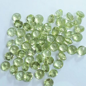 May include: A collection of light green peridot gemstones, each with a round, faceted shape. The gemstones are scattered on a white surface.