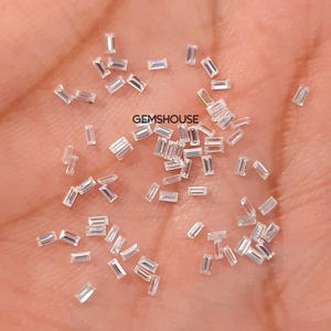 Può includere: Una collezione di piccole gemme rettangolari e trasparenti sparse su una mano. Le gemme sono sfaccettate, riflettono la luce e sembrano di alta qualità. Il testo "GEMSHOUSE" è visibile.