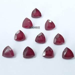May include: Ten triangular-cut, deep red gemstones are arranged on a white surface. The faceted surfaces of the gemstones reflect light, showcasing a rich, saturated colour. The text "GEMSHOUSE" is visible.