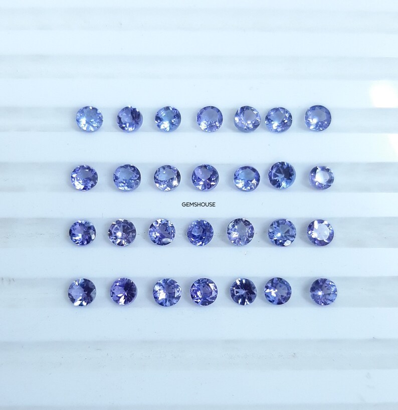 Pu&ograve; includere: Ventiquattro gemme rotonde, sfaccettate, di colore blu chiaro disposte in tre file da otto su una superficie bianca. GEMSHOUUSE &egrave; stampato in nero sotto le gemme.