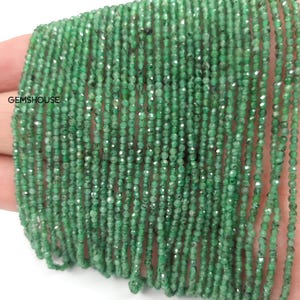 Può includere: Primo piano di fili di piccole perline verde smeraldo sfaccettate. Le perline sono infilate insieme, creando un aspetto fluido e strutturato. L'immagine include il testo "GEMSHOUSE".