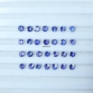 Pu&ograve; includere: Una collezione di 24 gemme rotonde, sfaccettate e blu disposte in quattro file di sei. Le gemme sono esposte su una superficie bianca con un sottile motivo a griglia. Il testo "GEMSHOUUSE" &egrave; visibile al centro dell'immagine.