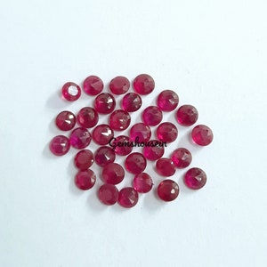 May include: A scattering of faceted, round, deep red gemstones on a white background. The gemstones have a polished, reflective surface. The image includes the text "gemshousein".