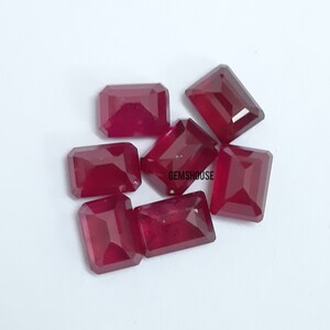 May include: Seven rectangular, faceted, deep red gemstones are displayed on a white surface. The gems are arranged in a cluster, with the text "GEMSHOUSE" visible on one of the gems. The gems have a polished, reflective surface.