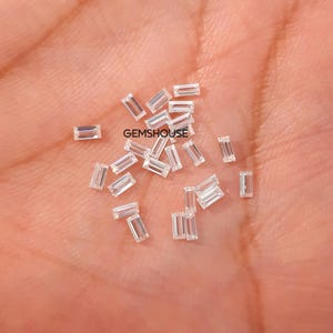 Può includere: Una collezione di piccole gemme rettangolari e trasparenti sparse su una superficie chiara. Le gemme sono sfaccettate, riflettendo la luce. Il testo "GEMSHOUSE" è visibile. Queste gemme sono adatte per la creazione di gioielli.