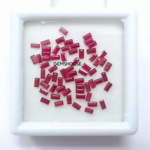 Puede incluir: Una colección de piedras preciosas de rubí rojo facetadas y de forma rectangular. Las piedras preciosas están dispuestas en una bandeja blanca con el texto "GEMSHouse" impreso en la bandeja.