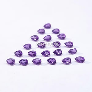 May include: A collection of 20 purple amethyst pear-shaped gemstones. The gemstones are arranged in a triangular pattern on a white surface.