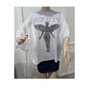 May include: A light gray t-shirt featuring the text NIRVANA in a distressed font above a dark gray graphic of a winged figure. The shirt has loose, flowing white short sleeves and a round neckline, paired with a solid dark blue mini skirt.