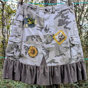 Puede incluir: Falda con estampado de camuflaje con parches de girasol, foto enmarcada y diseño floral. La falda tiene bolsillos de carga y un dobladillo con volantes. La falda está colgada al aire libre en un tendedero.